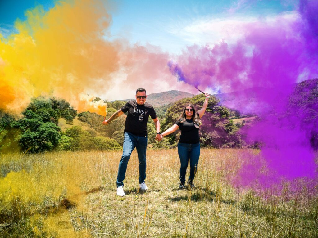 A joyful couple holding smoke bombs in a vibrant open field, celebrating together.